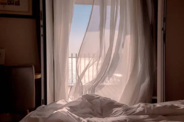 A cool breeze blowing light curtains through an open bedroom window.
