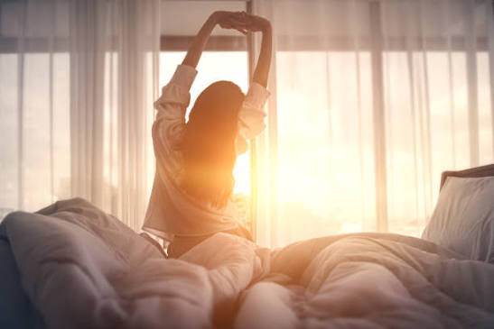 A woman stretching in bed as morning sun comes through the curtains.