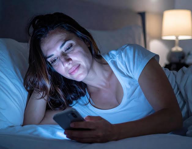 A tired woman lying in bed at night, looking at her phone screen.